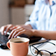 A keyboard being typed on in the distance. In the foreground is a coffee cup and reading glasses set down on the desktop.